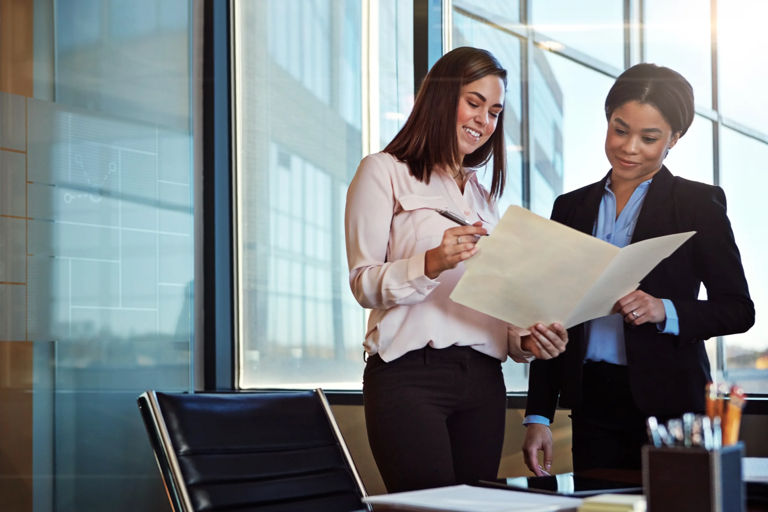 Two smiling professionals stand by a window in a modern office, examining a document folder and signing with a pen.