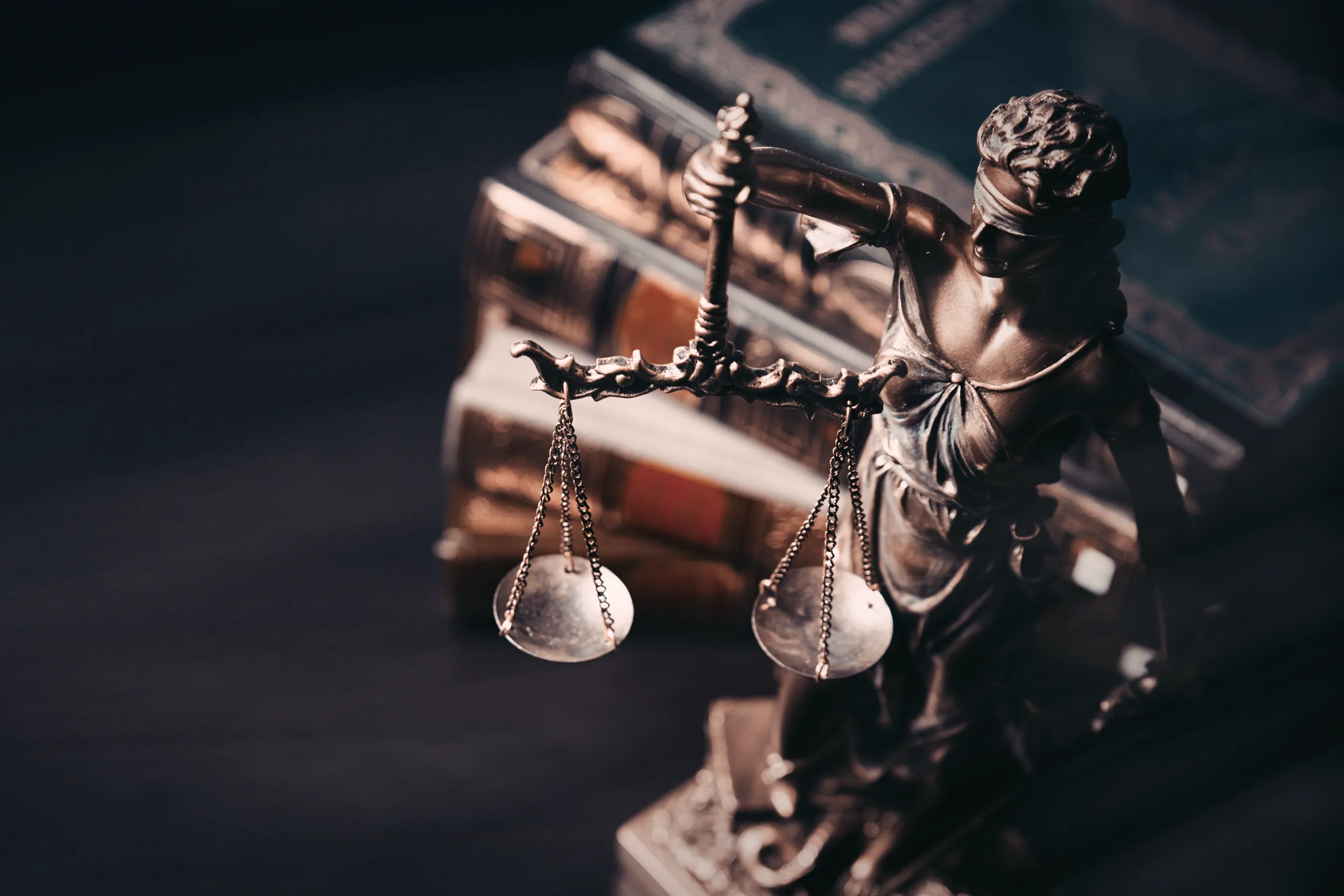A bronze statue of Lady Justice with balanced scales stands in front of a stack of blurred, dark leather-bound law books.