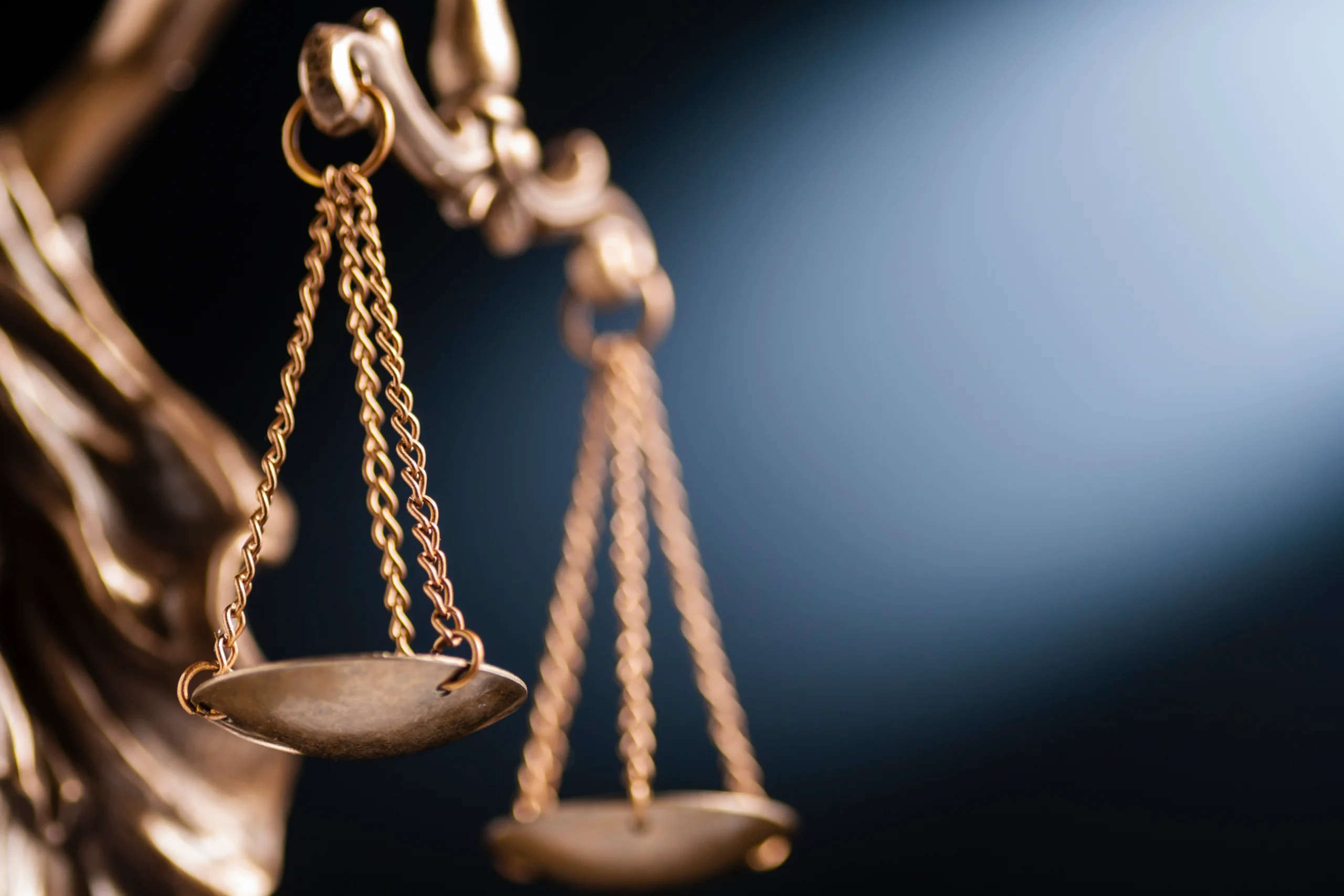A close-up of the brass scales of justice, suspended by chains against a dark background, dramatically highlighted by a beam of light.