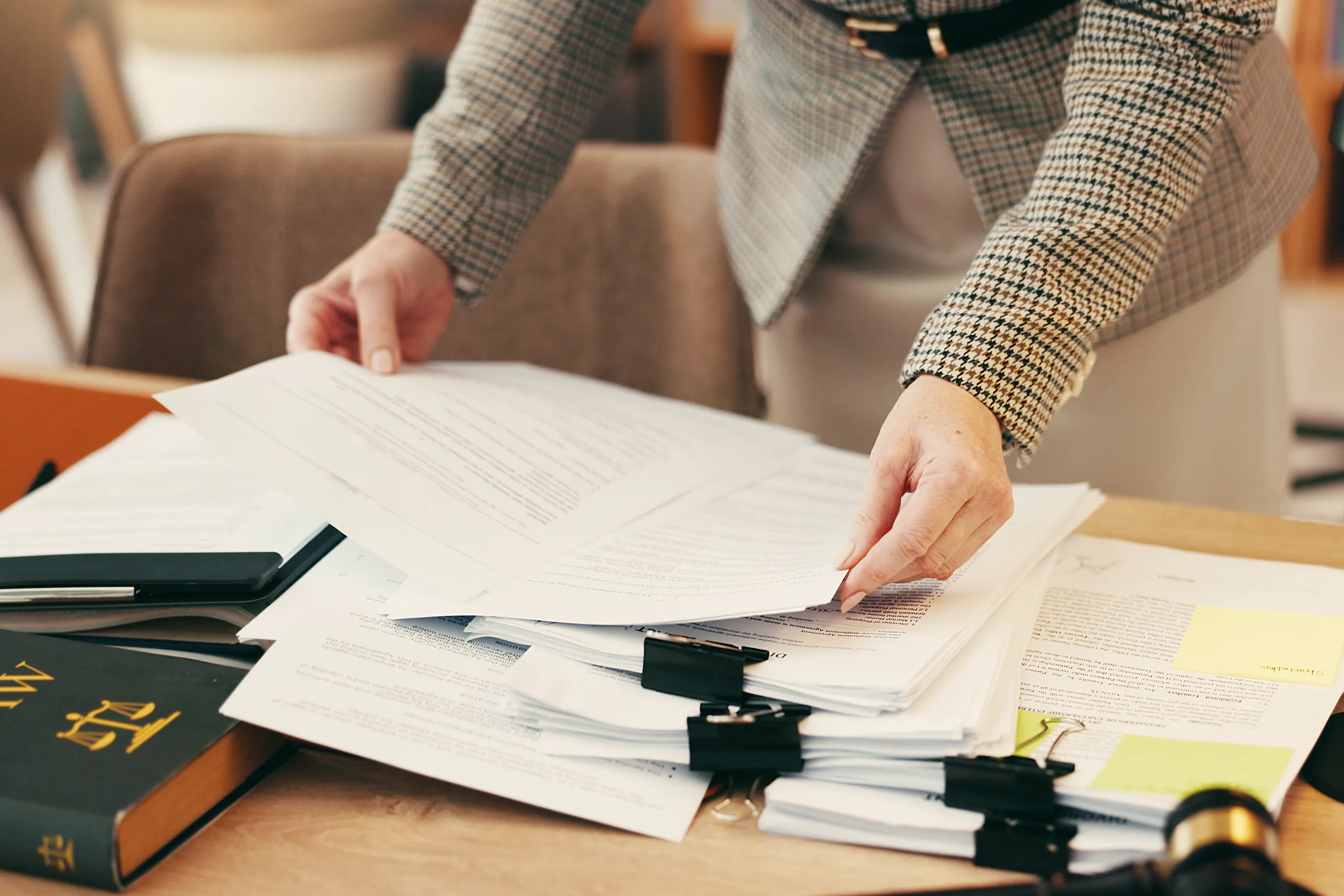 A person wearing a houndstooth blazer reviews stacks of clipped legal documents on a desk, next to a law book displaying the scales of justice.